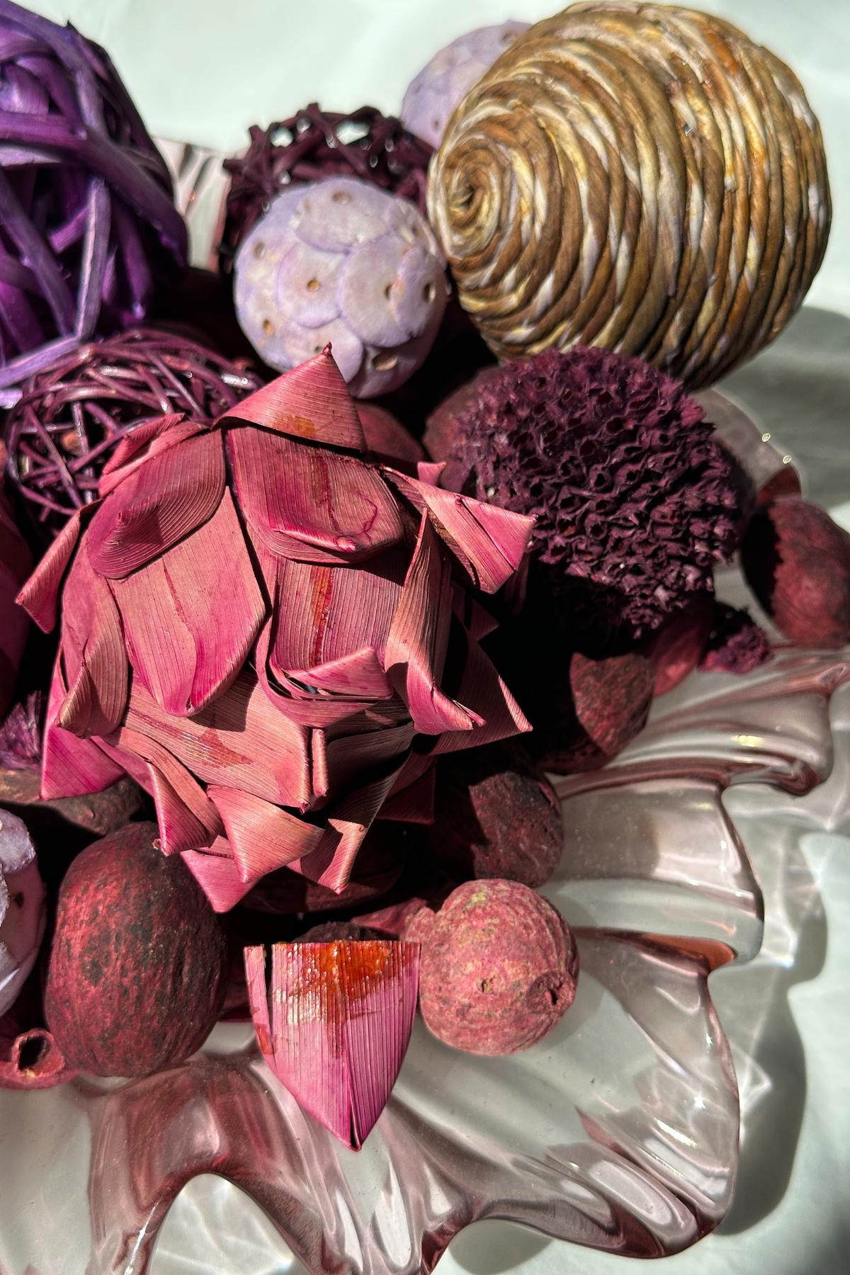 Close-up of chunky eco-friendly bowl fillers in shades of purple and pink, featuring dried flowers, woven natural spheres, and dried fruits arranged in a clear glass decorative bowl. Sustainable home decor accent.