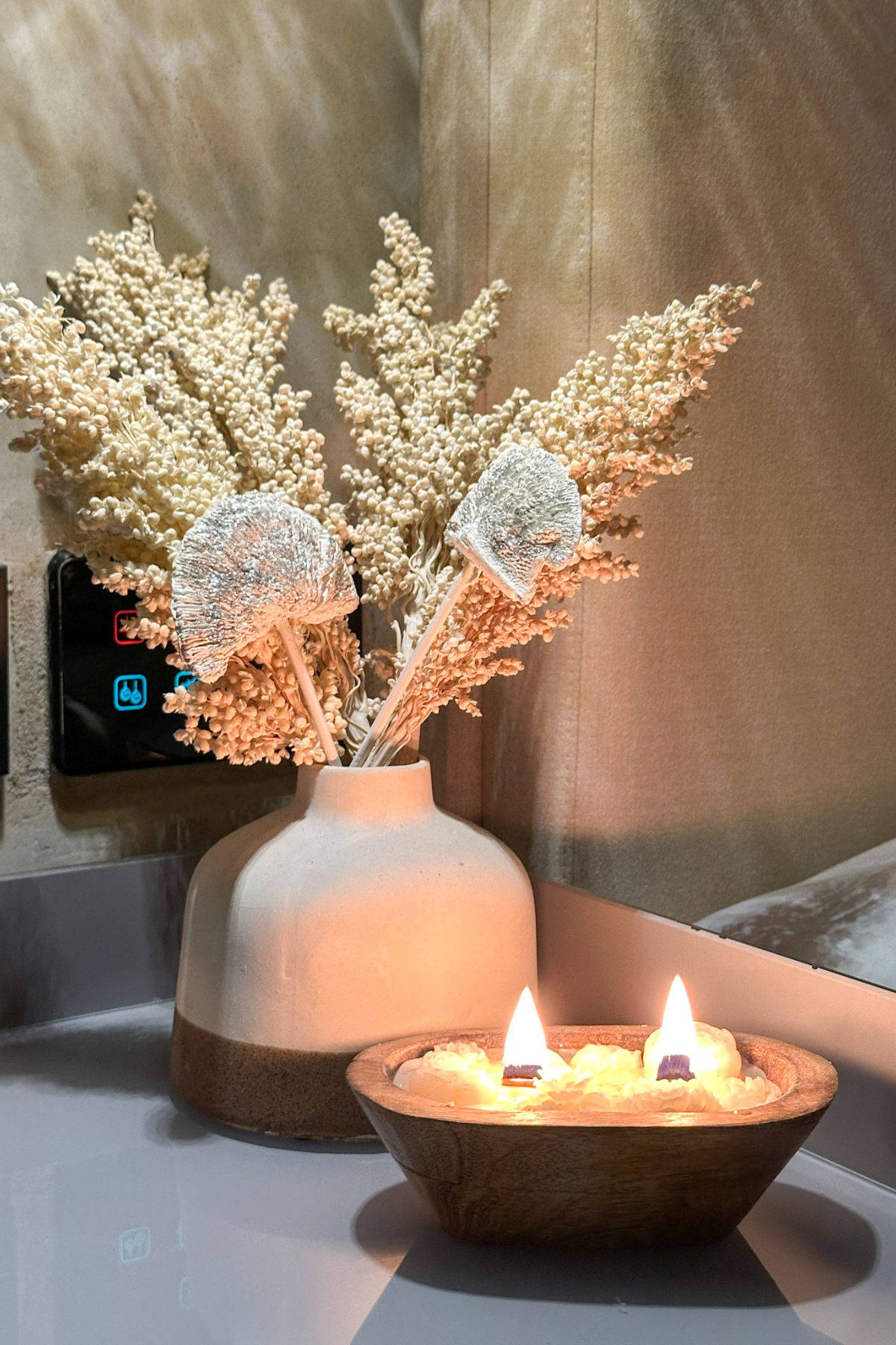 Decorative vase with dried flowers and two lit candles in a wooden bowl on a surface.