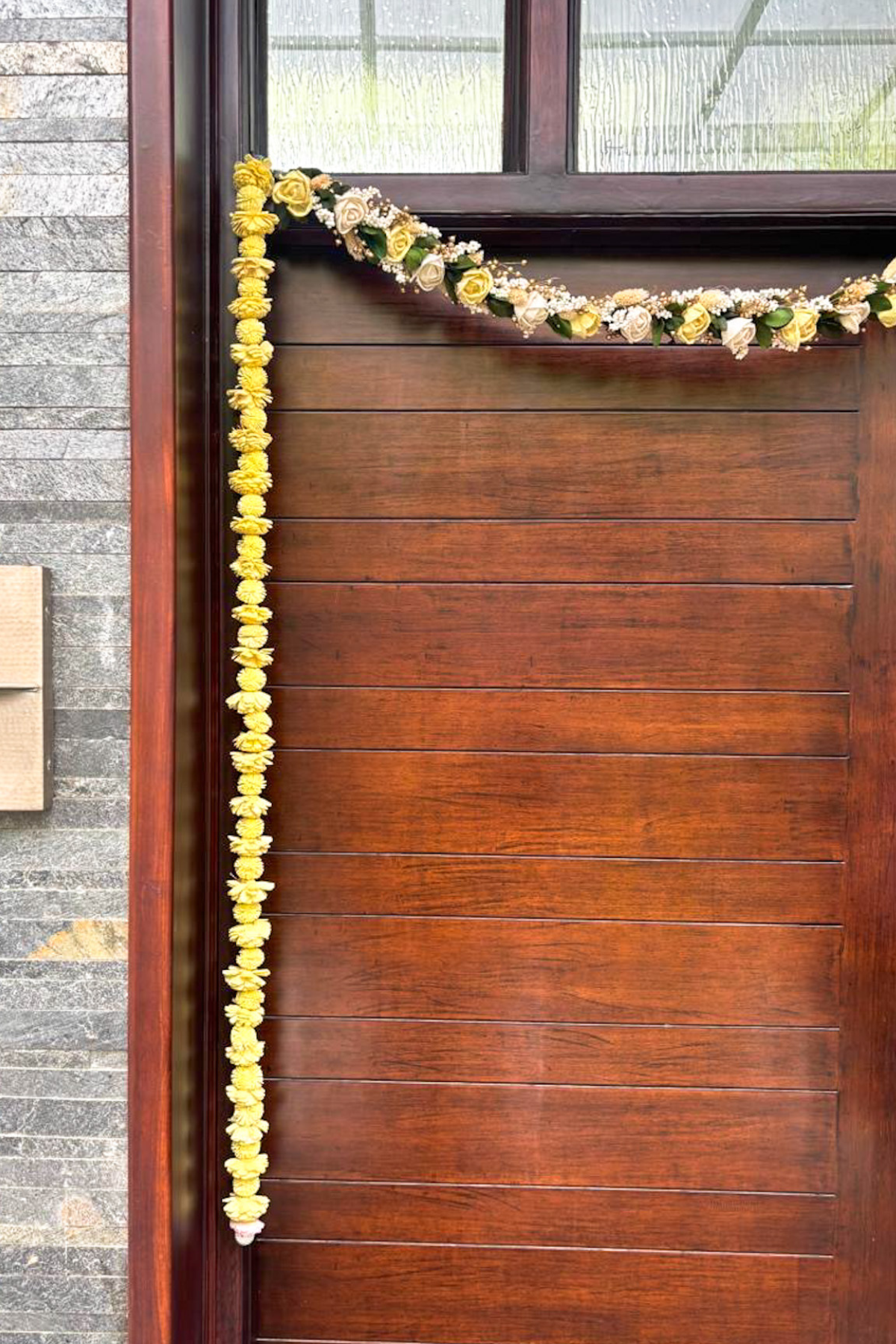 Vertical yellow flower toran garland hanging along a wooden front door, with a smaller floral garland draped across the top