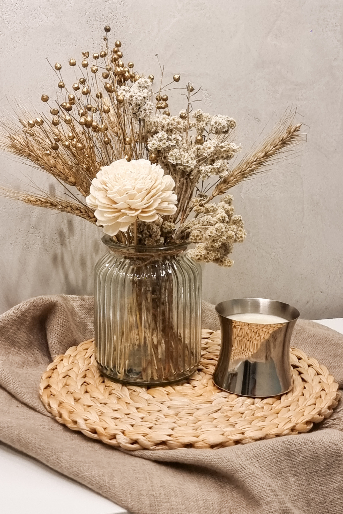 A dried floral bunch arrangement with daisy placed in a clear glass vase, with a metallic ribbed vase alongside, displayed on a woven mat. The arrangement includes shades of white, brown, and beige flowers.