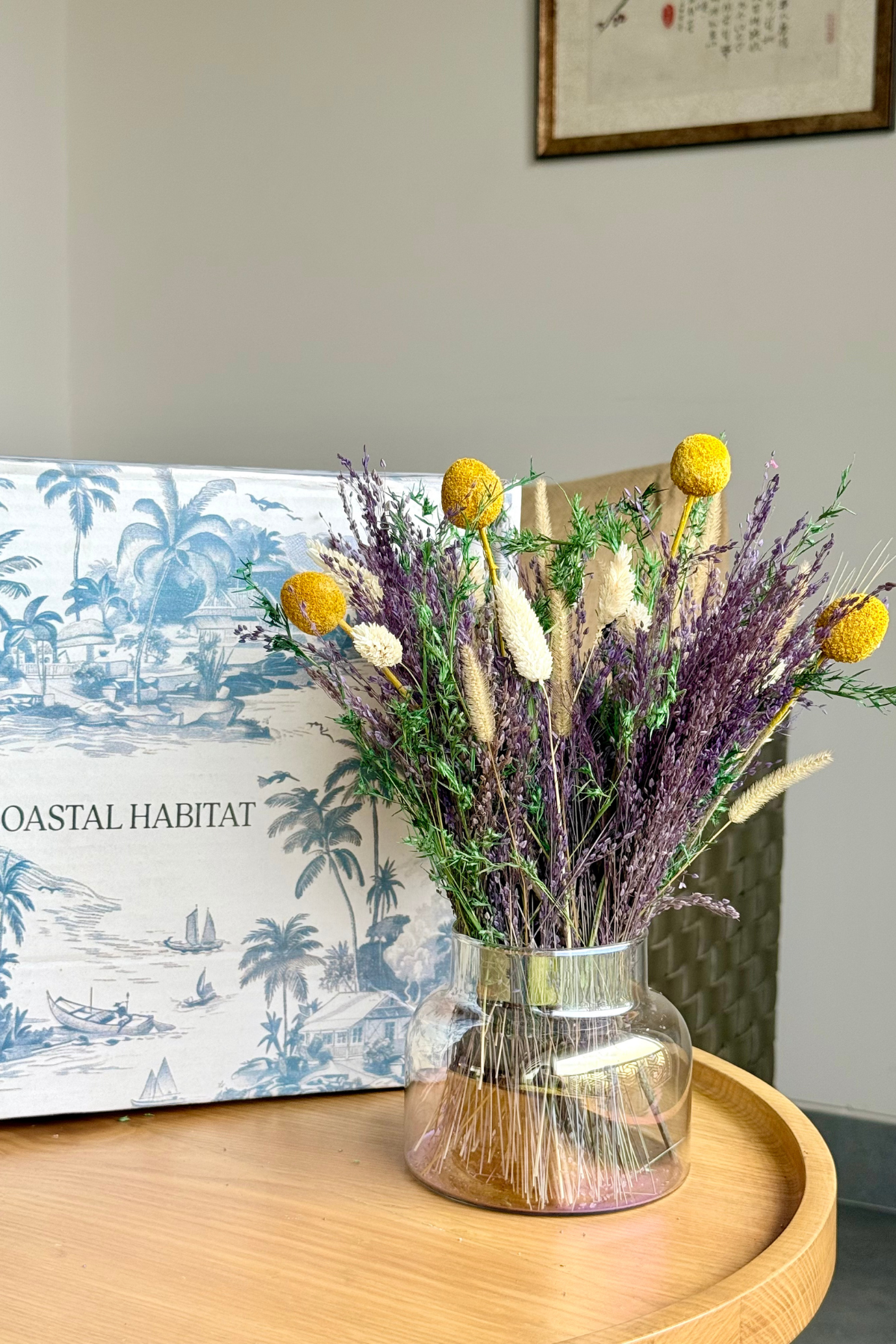 A glass jar vase filled with dried flowers, including purple lavender-like stems, yellow globe blooms, and beige grasses, placed on a round wooden tray. A decorative box with blue tropical artwork and the text 'COASTAL HABITAT' sits behind it against a light wall.